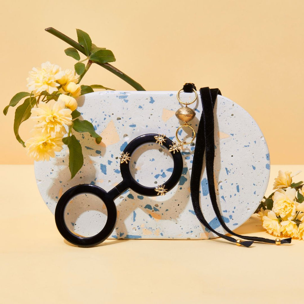 Colgante gafas Hormigas, realizado en acetato color negro con hormigas metálicas doradas y collar de terciopelo negro con fornituras  y abalorios dorados. El colgante tiene una bisagra que hace que se pueda desplegar y transformar en gafas de lectura.