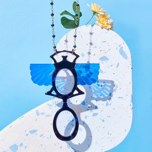 Colgante-gafas en forma de escarabajo egipcio. Realizado en combinación de acetato imitación carey en tonos azules con las alas en azul klein transparente. Cadena de rosario en latón con cuentas de vidrio azul. Una bisagra hace que se pueda transformar en gafas de lectura