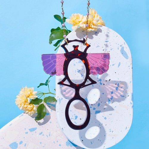 Colgante-gafas en forma de escarabajo egipcio. Realizado en combinación de acetato imitación carey con las alas en rosa transparente. Cadena de rosario en latón con cuentas de vidrio rosa. Una bisagra hace que se pueda transformar en gafas de lectura