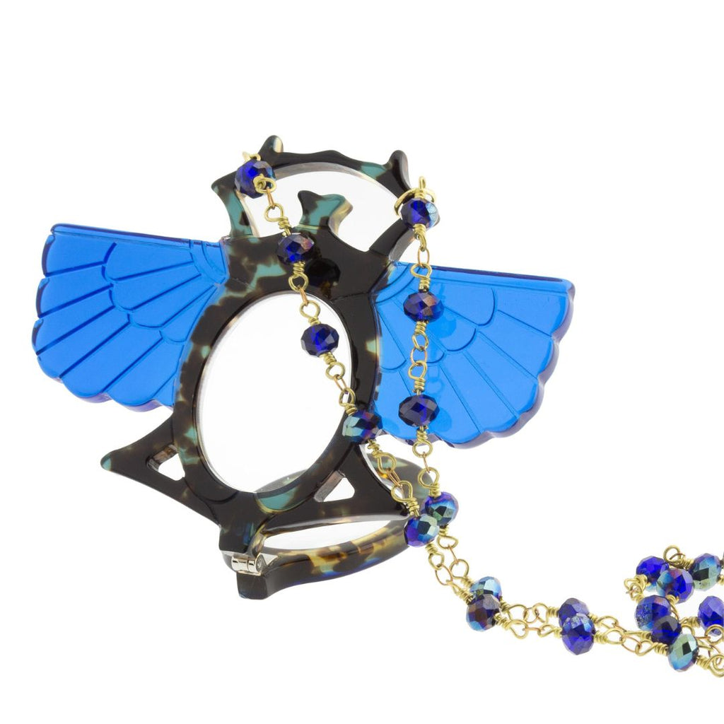 Colgante-gafas en forma de escarabajo egipcio. Realizado en combinación de acetato imitación carey en tonos azules con las alas en azul klein transparente. Cadena de rosario en latón con cuentas de vidrio azul. Una bisagra hace que se pueda transformar en gafas de lectura