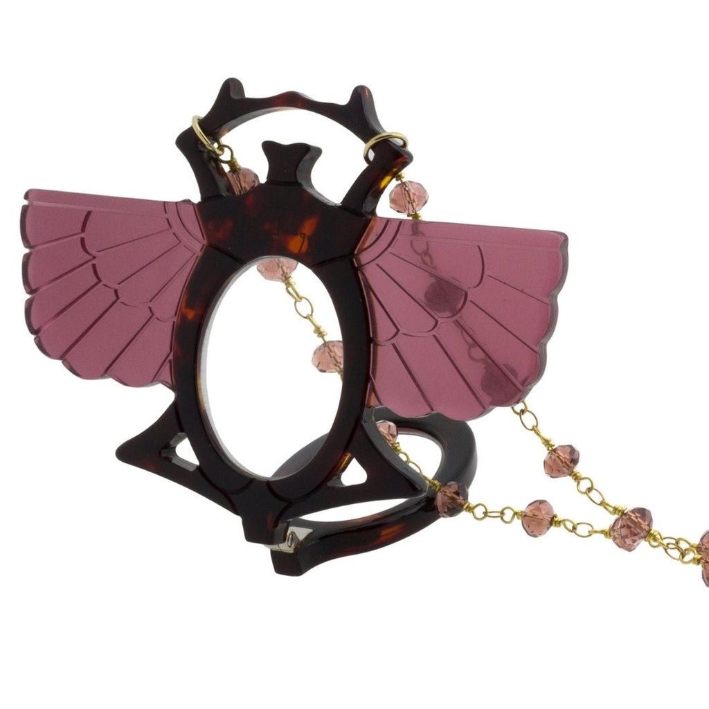 Colgante-gafas en forma de escarabajo egipcio. Realizado en combinación de acetato imitación carey con las alas en rosa transparente. Cadena de rosario en latón con cuentas de vidrio rosa. Una bisagra hace que se pueda transformar en gafas de lectura