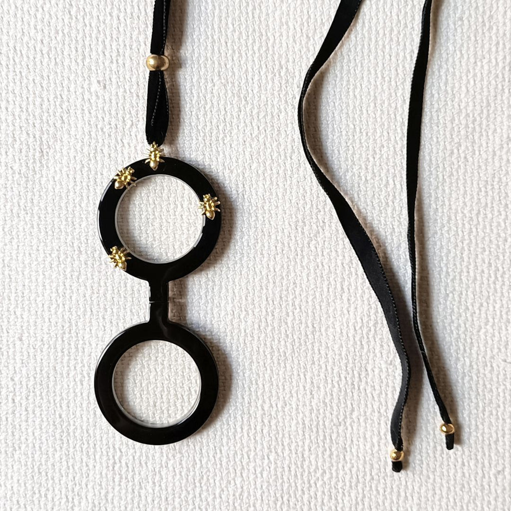 Colgante gafas Hormigas, realizado en acetato color negro con hormigas metálicas doradas y collar de terciopelo negro con fornituras  y abalorios dorados. El colgante tiene una bisagra que hace que se pueda desplegar y transformar en gafas de lectura.