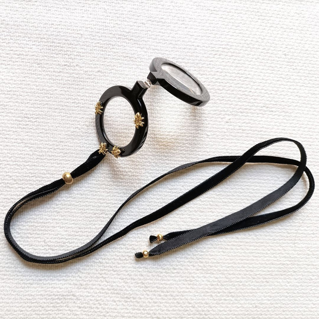 Colgante gafas Hormigas, realizado en acetato color negro con hormigas metálicas doradas y collar de terciopelo negro con fornituras  y abalorios dorados. El colgante tiene una bisagra que hace que se pueda desplegar y transformar en gafas de lectura.