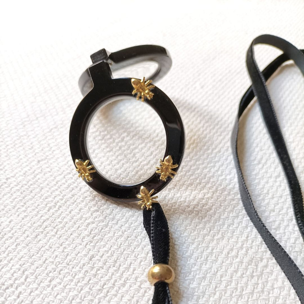 Colgante gafas Hormigas, realizado en acetato color negro con hormigas metálicas doradas y collar de terciopelo negro con fornituras  y abalorios dorados. El colgante tiene una bisagra que hace que se pueda desplegar y transformar en gafas de lectura.