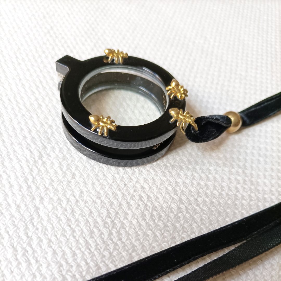 Colgante gafas Hormigas, realizado en acetato color negro con hormigas metálicas doradas y collar de terciopelo negro con fornituras  y abalorios dorados. El colgante tiene una bisagra que hace que se pueda desplegar y transformar en gafas de lectura.