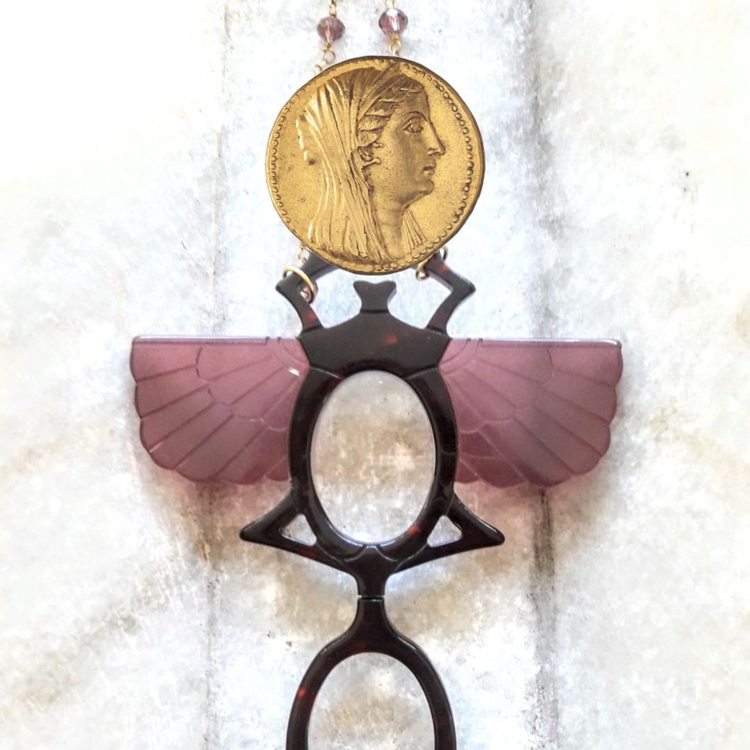 Colgante-gafas en forma de escarabajo egipcio. Realizado en combinación de acetato imitación carey con las alas en rosa transparente. Cadena de rosario en latón con cuentas de vidrio rosa. Una bisagra hace que se pueda transformar en gafas de lectura